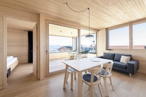 A modern interior with a wooden floor, a dining table with chairs, a sofa, and large windows offering a view of a mountain and a building.