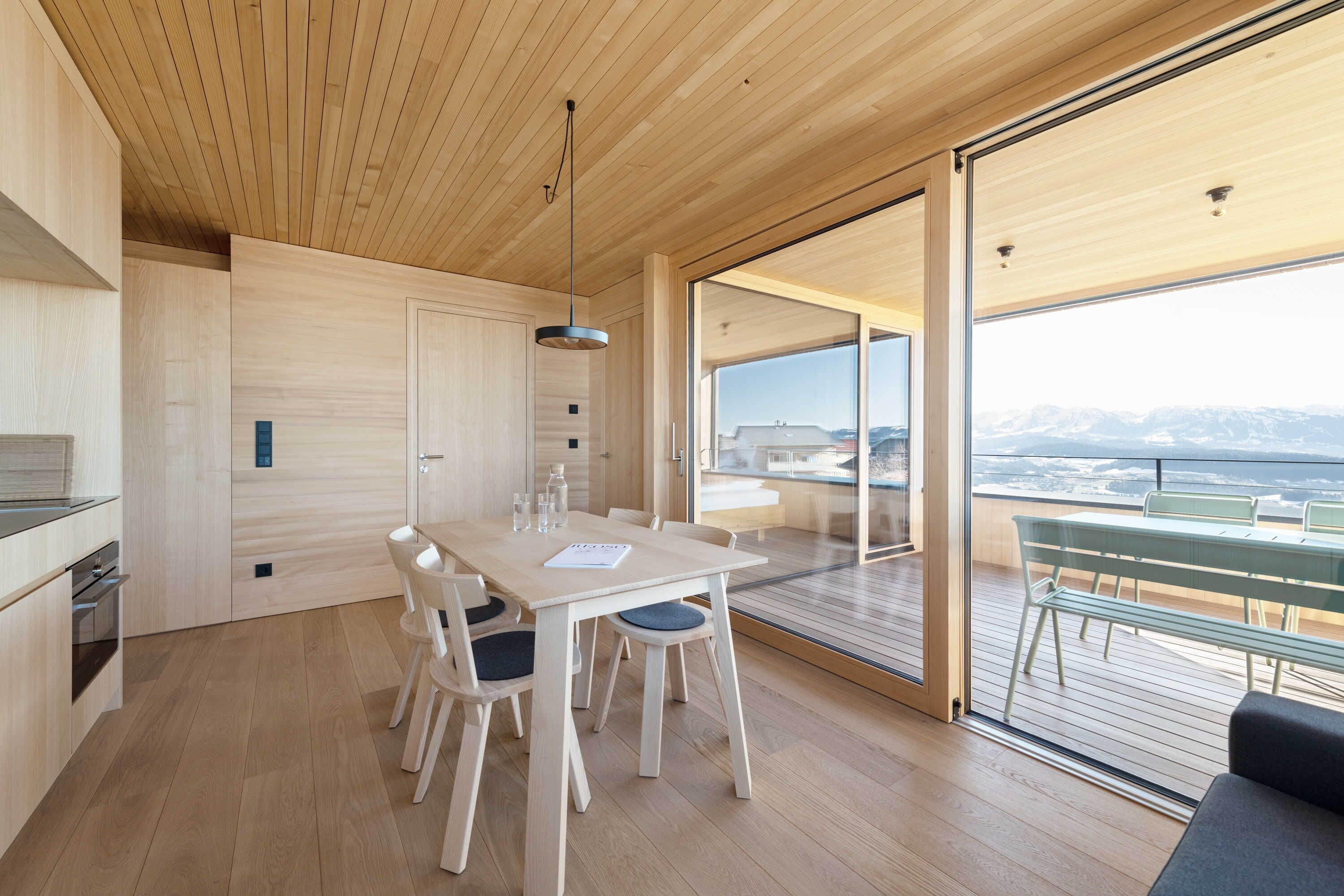 A modern kitchen with wooden cabinets, a dining table with chairs, and a view of the outdoors through large glass doors.