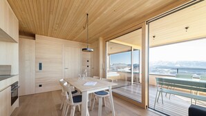 Een moderne keuken met houten kastjes, een eettafel met stoelen en uitzicht op het groen buiten via grote glazen deuren.