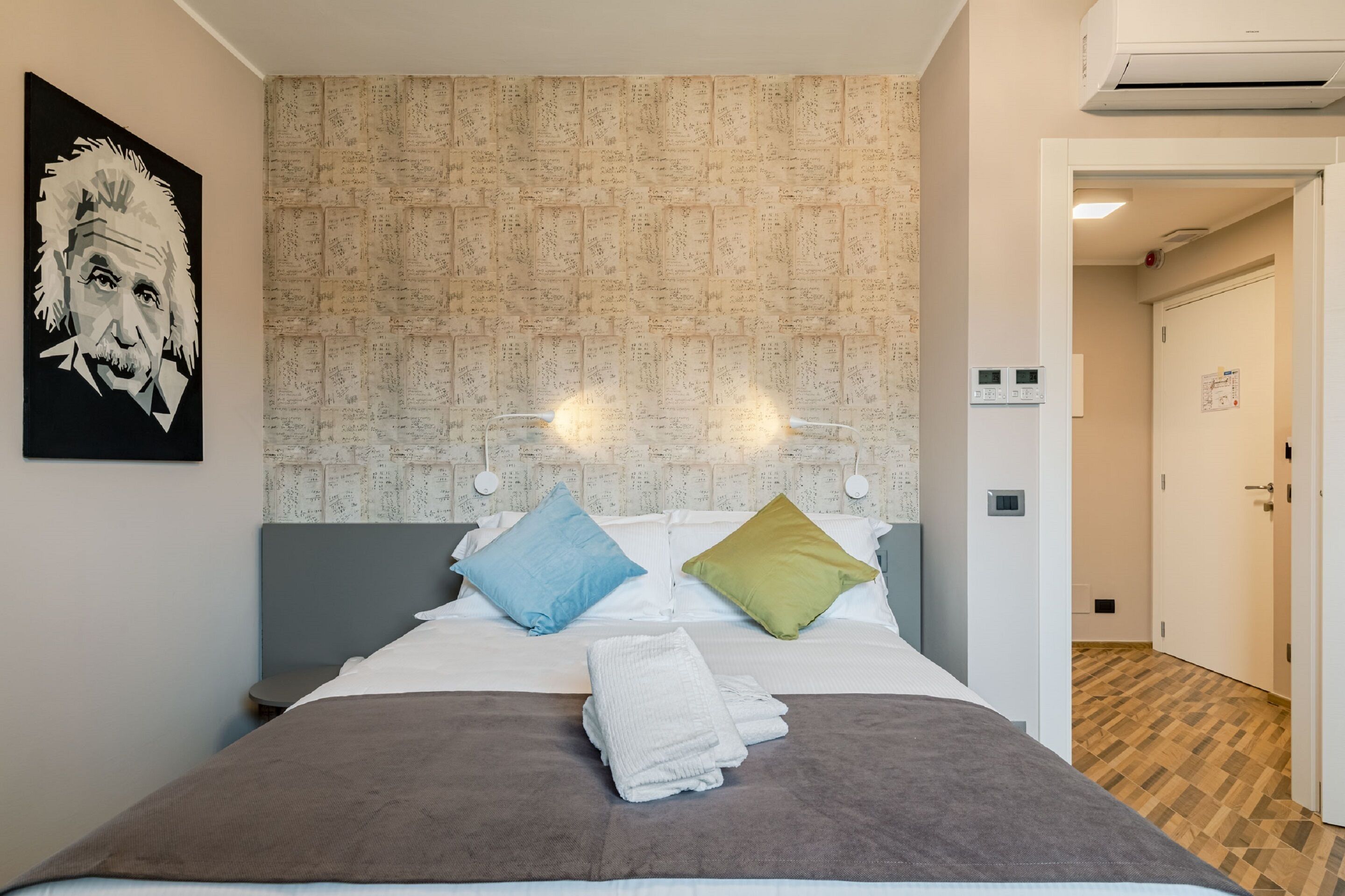 Ein modernes Hotelzimmer mit einem gemusterten Kopfteil, einem Bett mit zwei Kissen, einem ordentlich gefalteten weißen Handtuch und einem gerahmten Kunstwerk an der Wand.