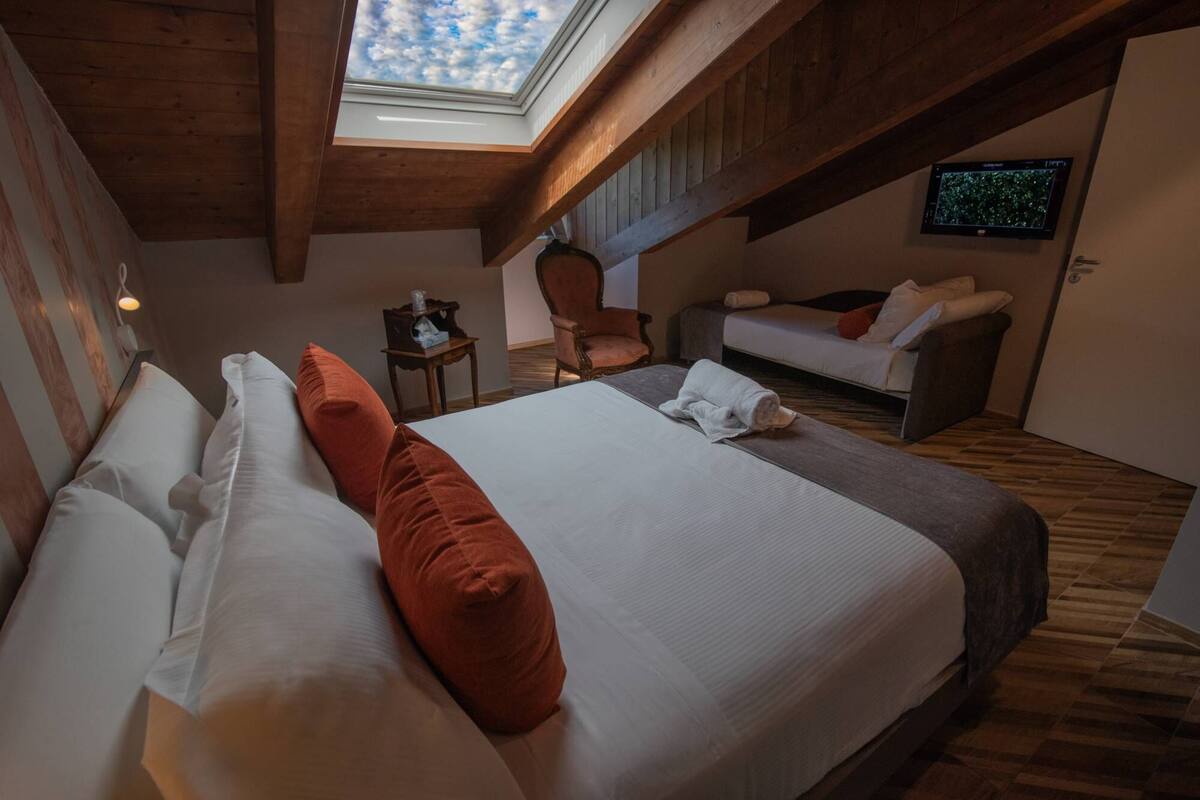 Un dormitorio con una cama grande, techo de madera, tragaluz, televisor en la pared y un sofá.