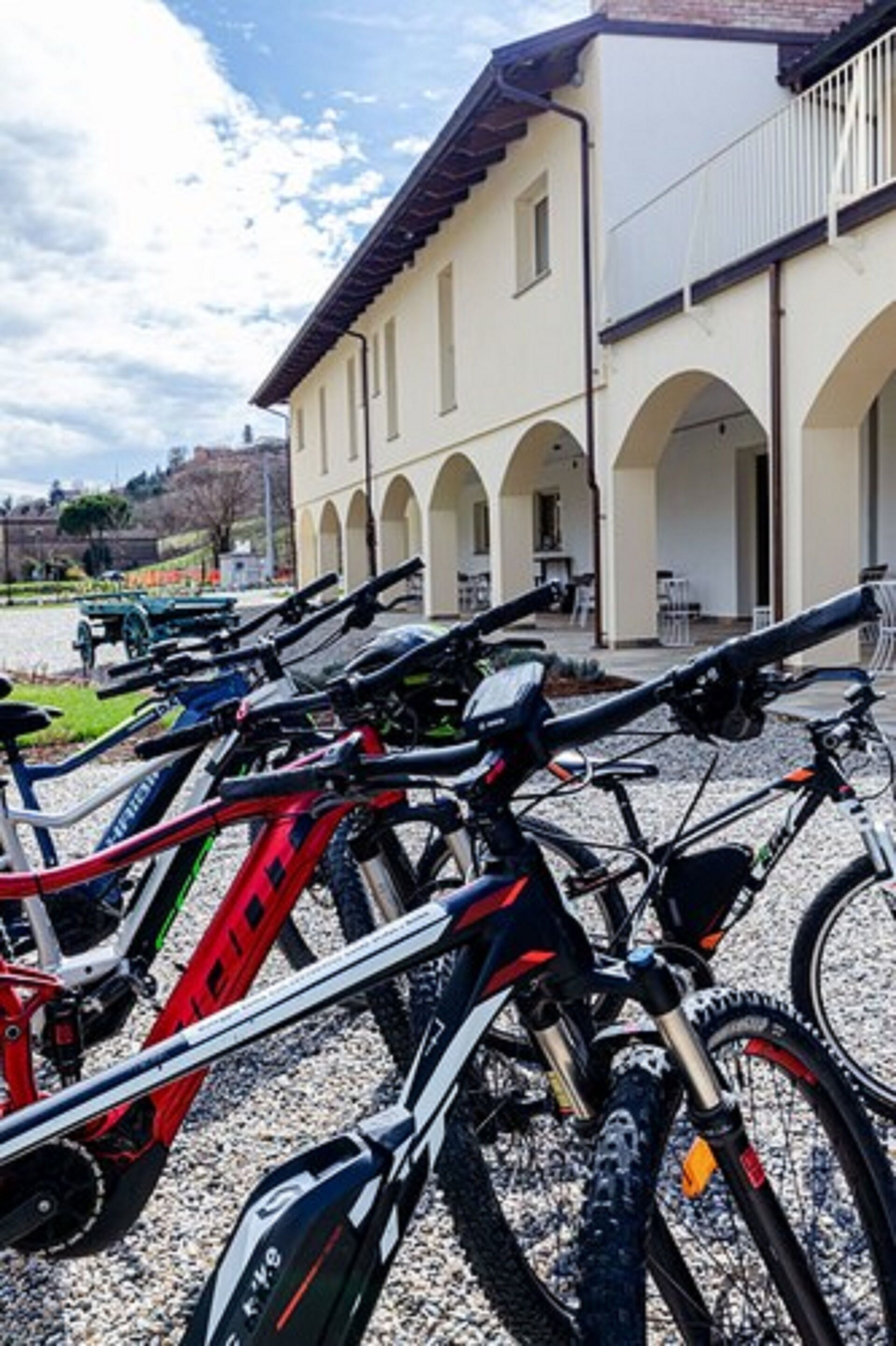 A row of bicycles parked in front of a building with arches and a gravel ground.