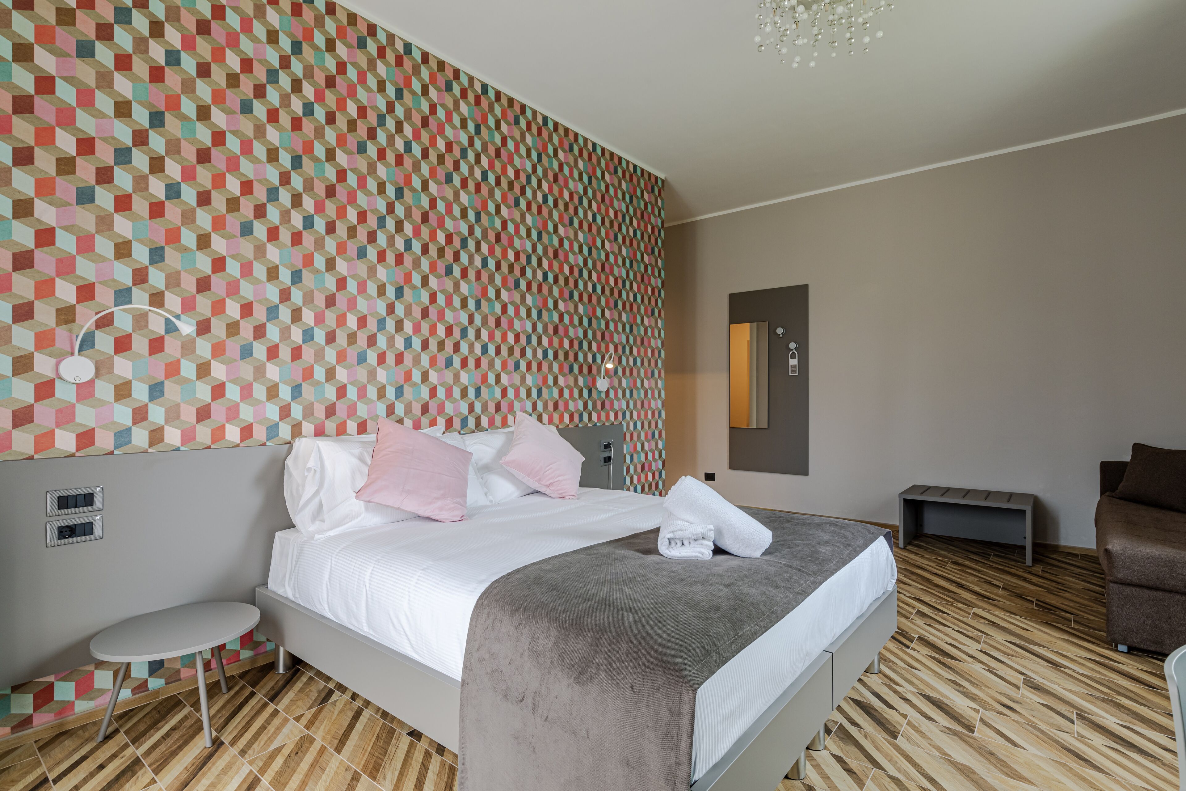 A modern hotel room with a geometric patterned wall, a neatly made bed with a grey blanket, a small round table, and a sofa.