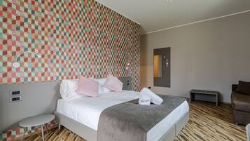 Una habitación de hotel moderna con un muro de diseño geométrico, una cama bien tendida con manta gris, una pequeña mesa redonda y un sofá.