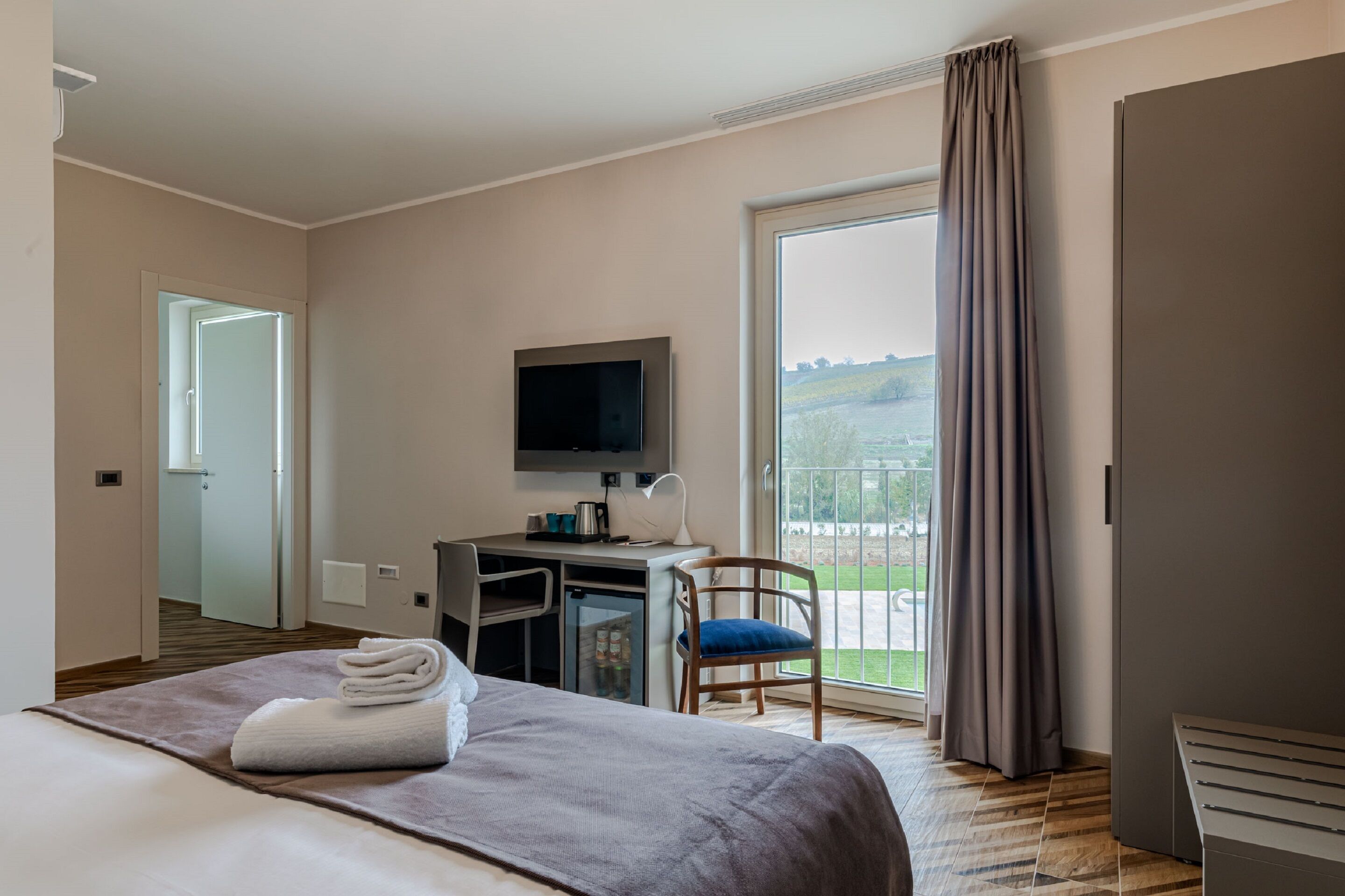 Ein Hotelzimmer mit Bett, Schreibtisch, Stuhl, Fernseher und Blick auf eine grüne Landschaft durch ein großes Fenster.