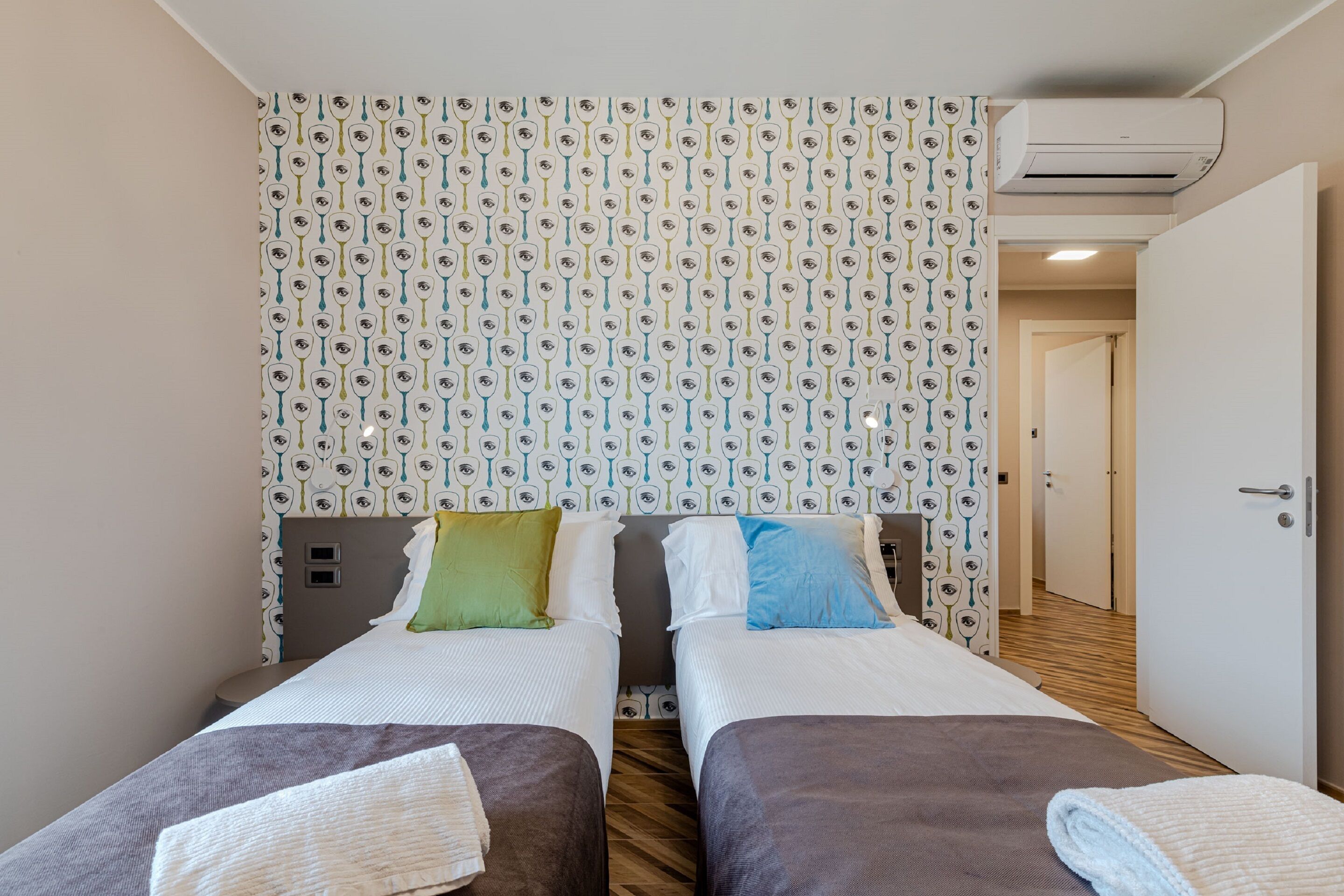 Habitación de hotel con dos camas, papel tapiz estampado, unidad de aire acondicionado y suelo de madera.