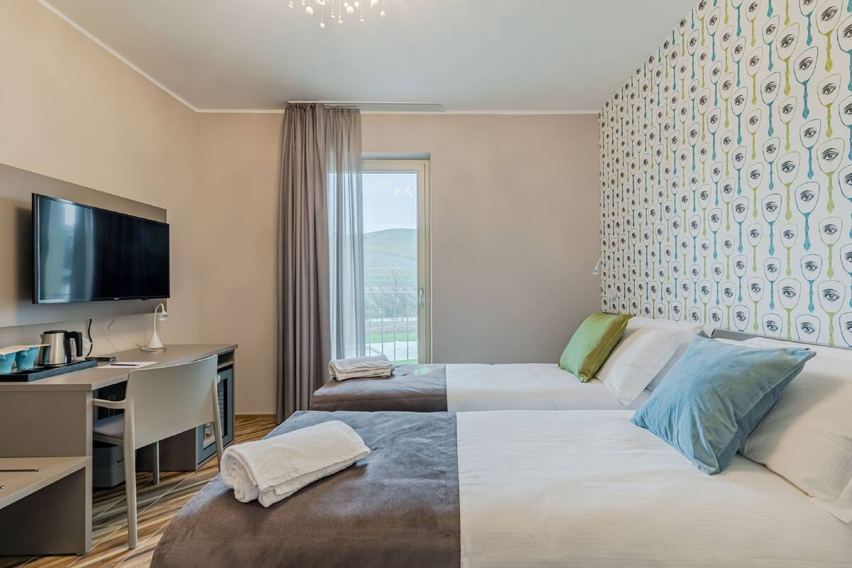 Habitación de hotel con televisión de pantalla plana, escritorio con lámpara, dos camas con almohadas, ventana con cortinas y una pared con diseño.