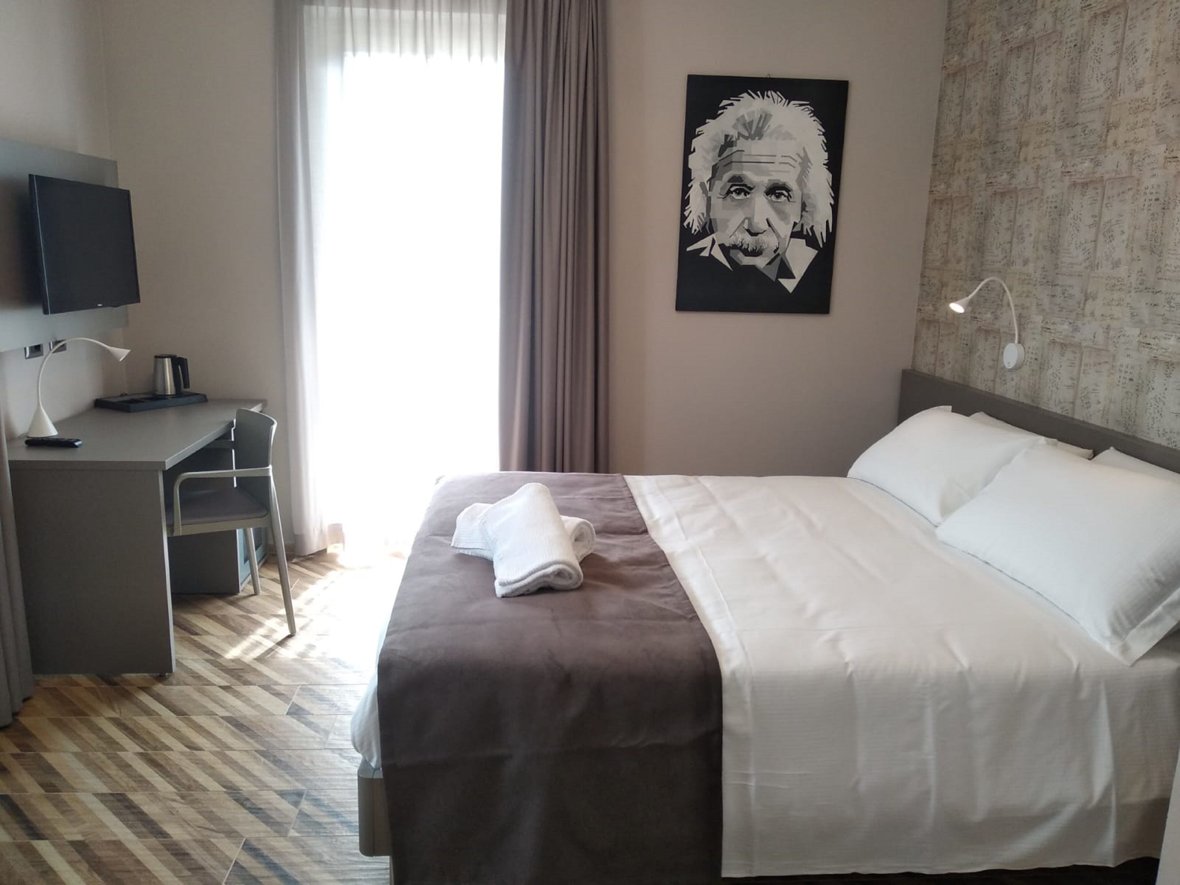 A hotel room with a bed, a desk, a chair, a television, and a poster on the wall.