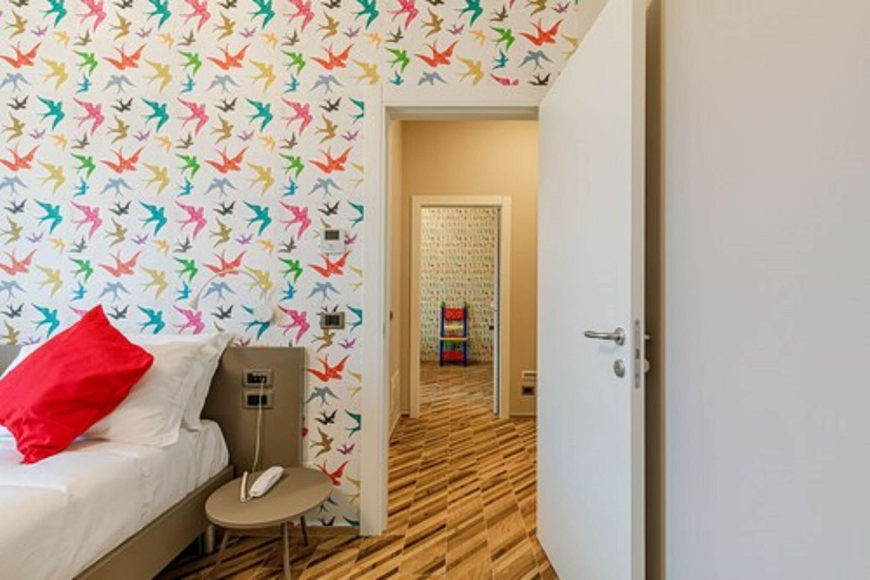 Ein Hotelzimmer mit Bett, Schreibtisch und Stuhl. Der Raum hat einen Holzboden und eine Wand mit bunten Vogelmotiven.