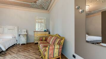 Une chambre d’hôtel avec un parquet en bois, un lit recouvert de draps blancs, un canapé à rayures, une commode et un grand miroir au mur.