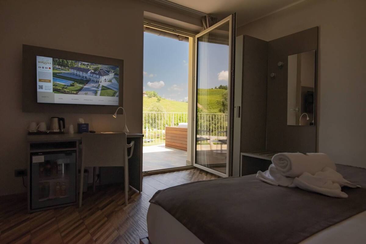 Habitación de hotel con cama, escritorio, televisor y balcón con vistas al campo.