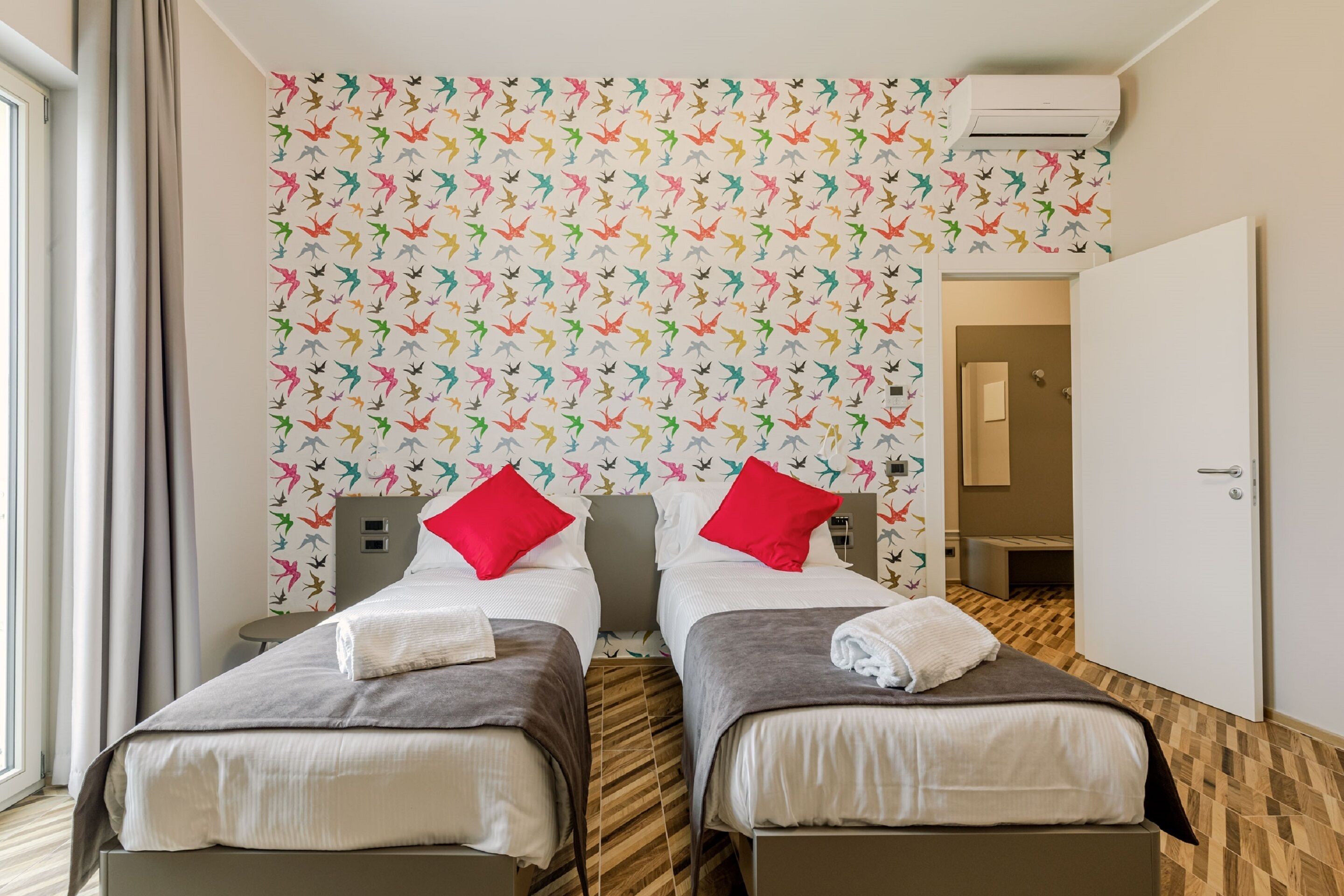 A hotel room with two beds, each with a pillow and a towel. The room has a patterned wallpaper and a wooden floor.