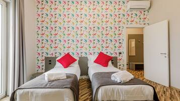 A hotel room with two beds, each with a pillow and a towel. The room has a patterned wallpaper and a wooden floor.
