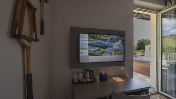 Quarto de hotel com TV fixada exibindo uma imagem do empreendimento, escrivaninha com cafeteira e janela com vista para o exterior.