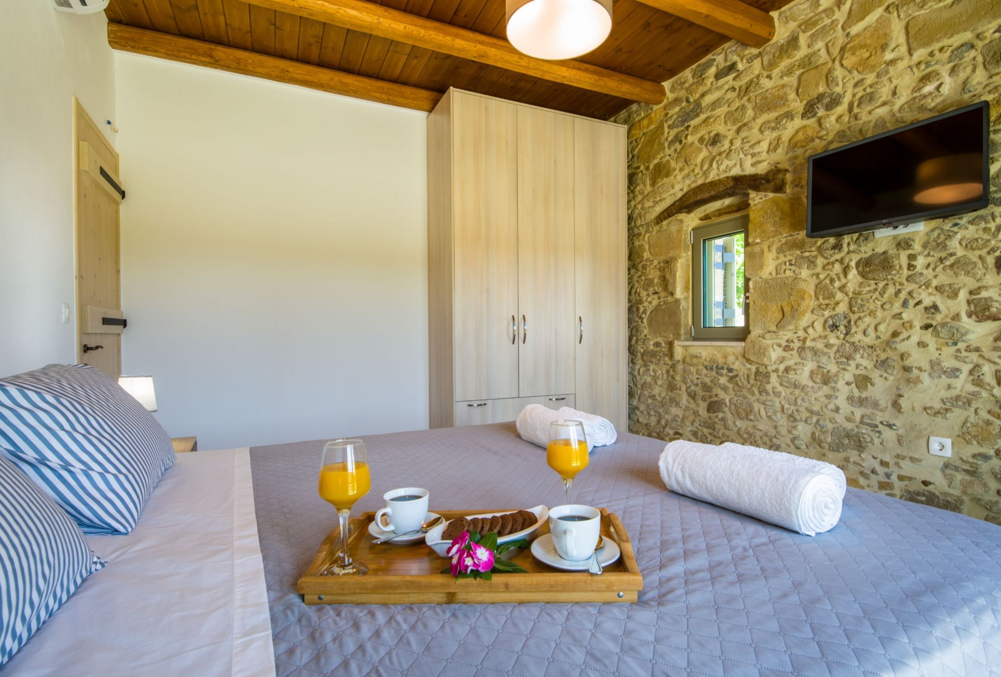 Une chambre avec un lit, un plateau en bois contenant des articles pour le petit-déjeuner, un mur en pierre et un téléviseur fixé au mur.
