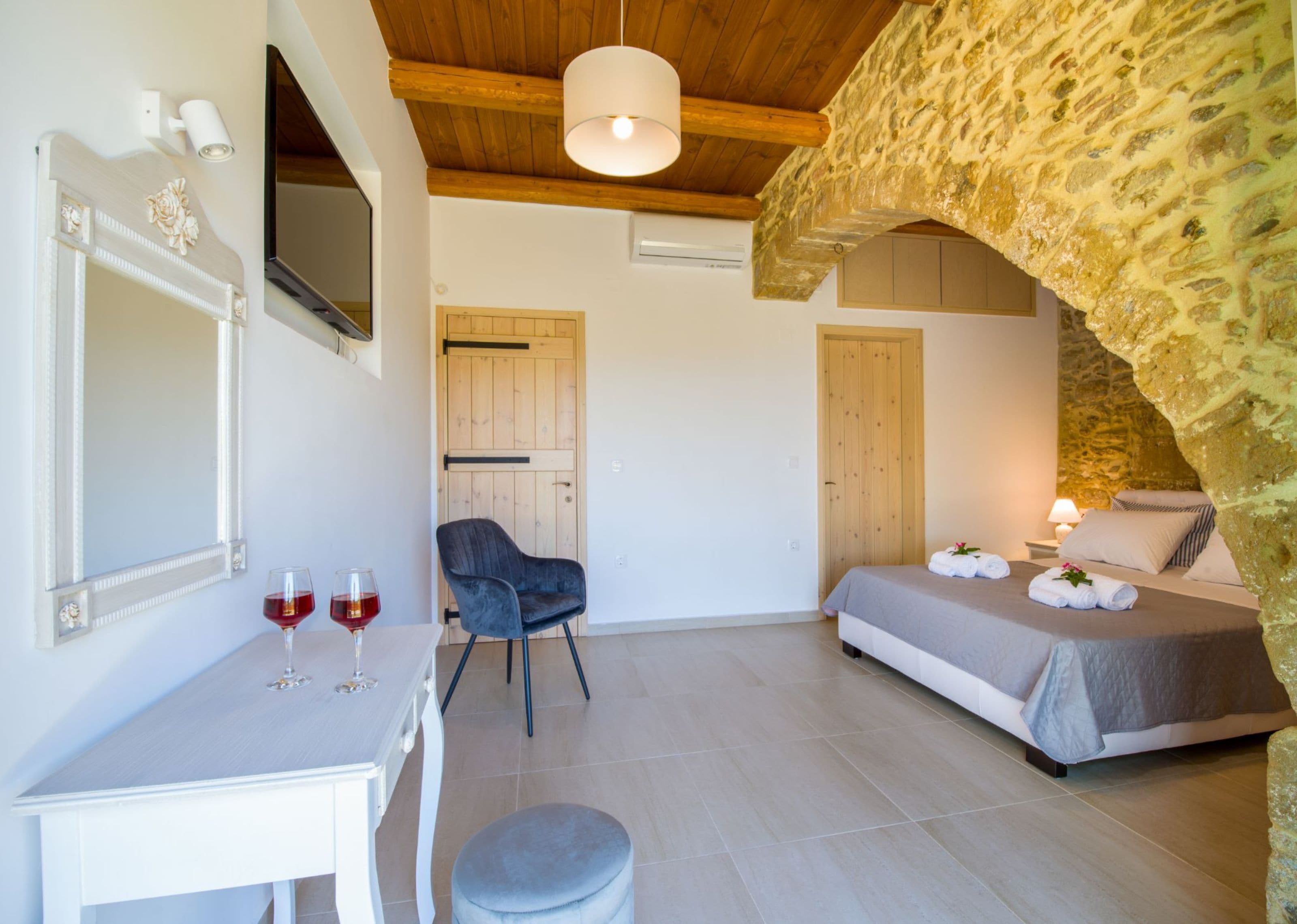 A bedroom with a stone wall, a bed with towels, a chair, a desk with a wine glass, and a mirror.