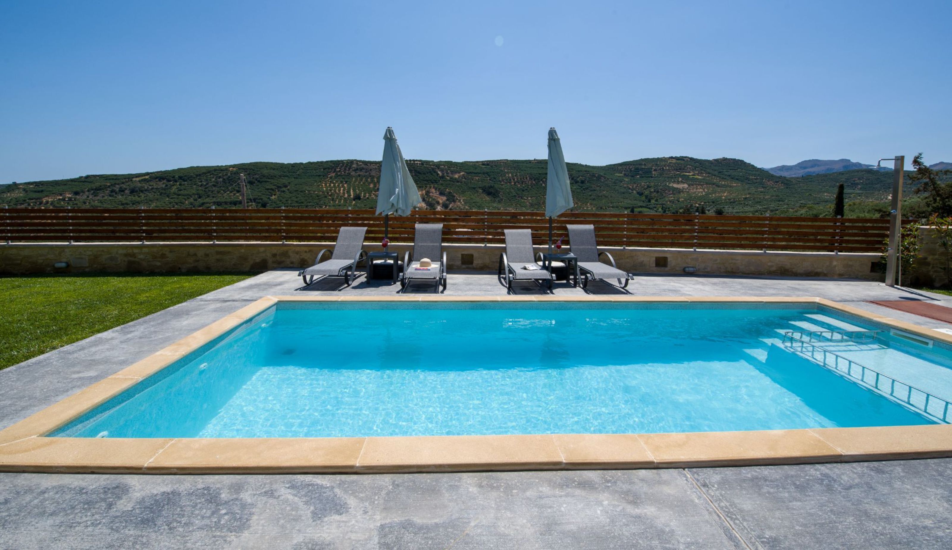 A rectangular swimming pool with a clear blue water, surrounded by a low stone wall. There are four lounge chairs with grey cushions and white umbrellas, and a wooden fence in the background.