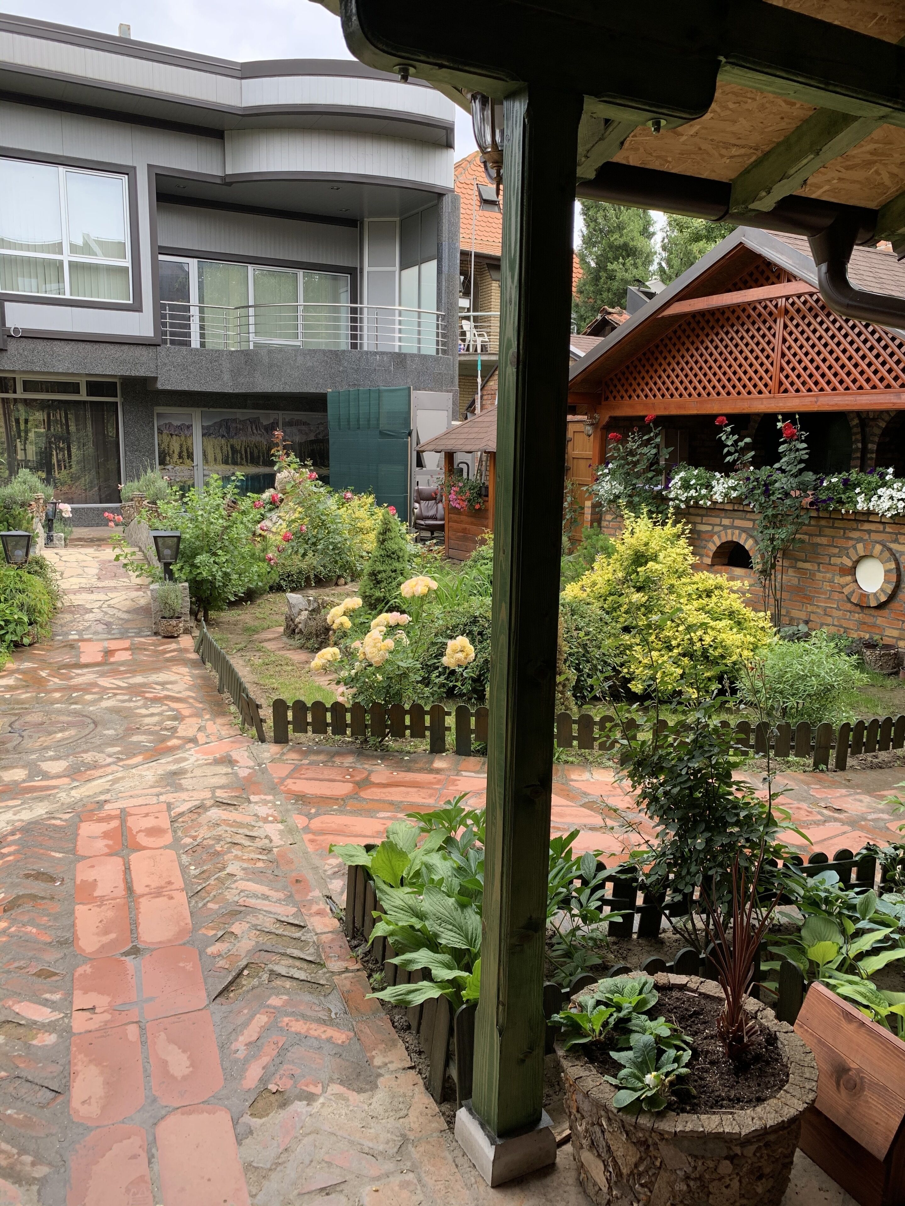 A courtyard with a stone pathway leading to a modern building with large windows and a balcony. There are various plants and flowers along the pathway, and a wooden structure with a lattice roof in the background.