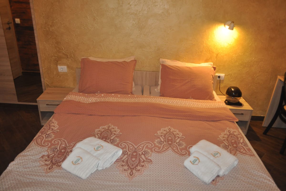 A neatly made bed with a patterned bedspread and pillows, two folded towels, a bedside lamp, and a nightstand with a lamp.