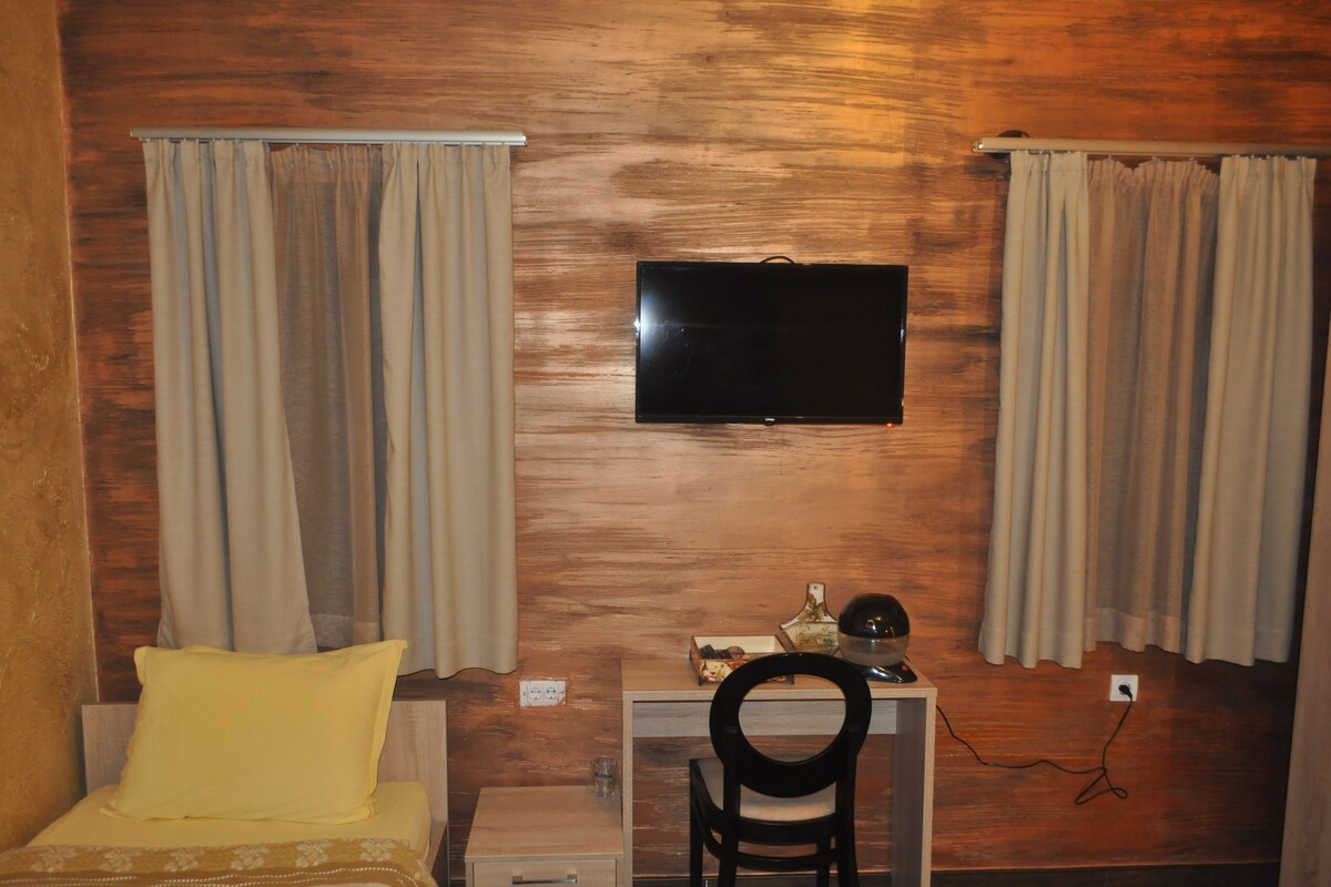 A room with a bed, a desk, a chair, a television mounted on the wall, and curtains on the windows.