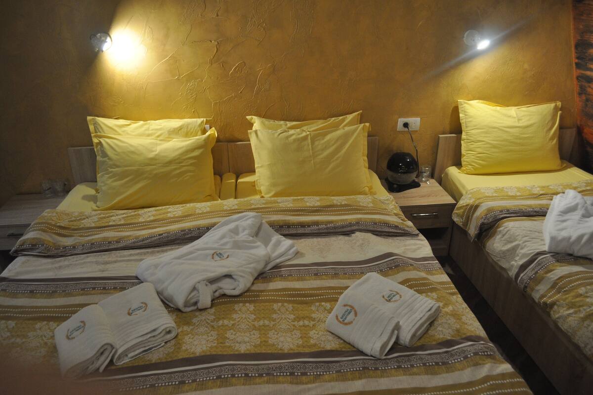 A hotel room with two beds, each with yellow pillows and striped bedding. There are towels on the beds and a nightstand with a lamp.