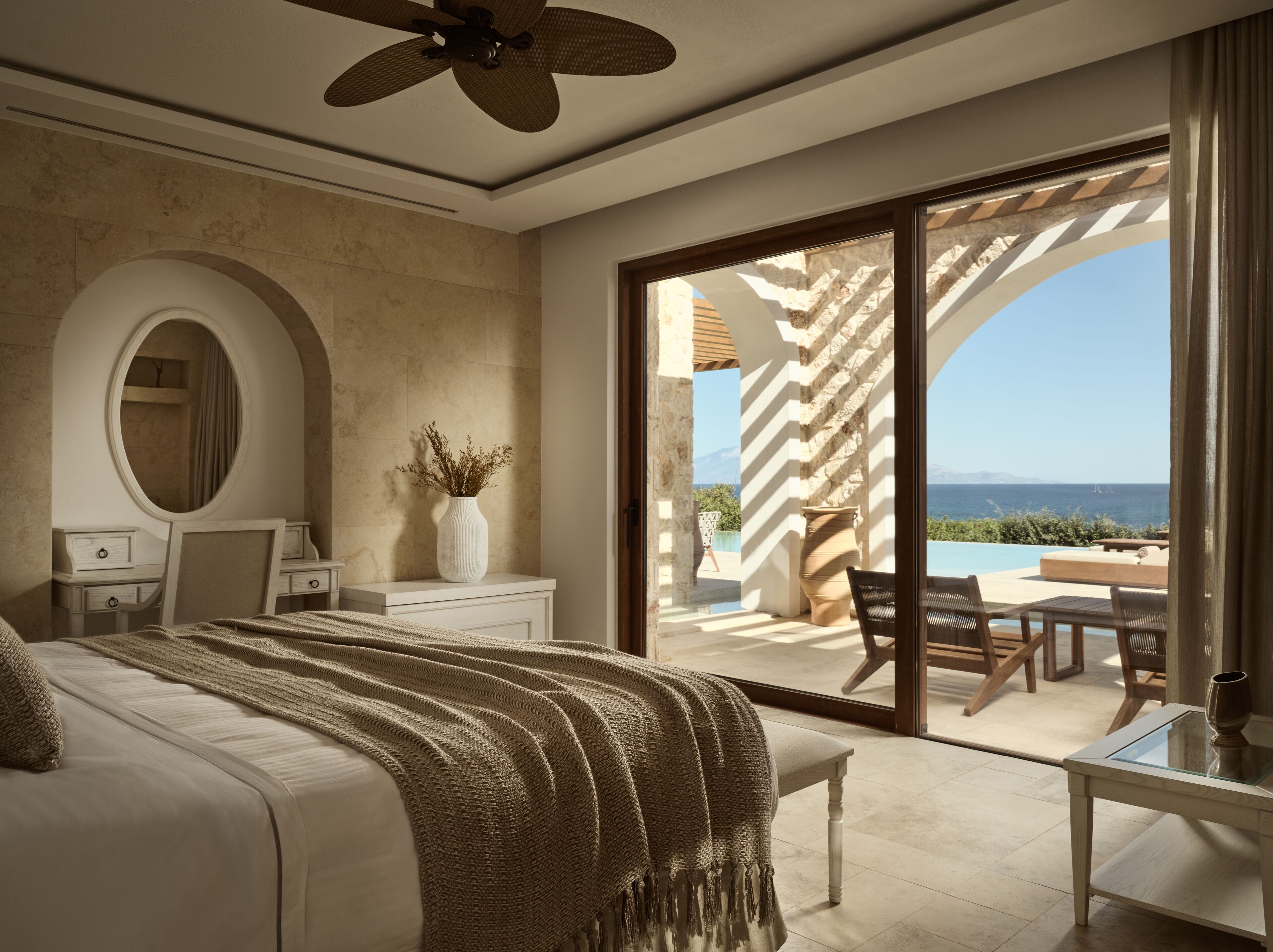 Une chambre dotée d’un grand lit, d’une coiffeuse avec un miroir, d’un vase contenant des plantes séchées, et offrant une vue sur une piscine extérieure et un espace patio.
