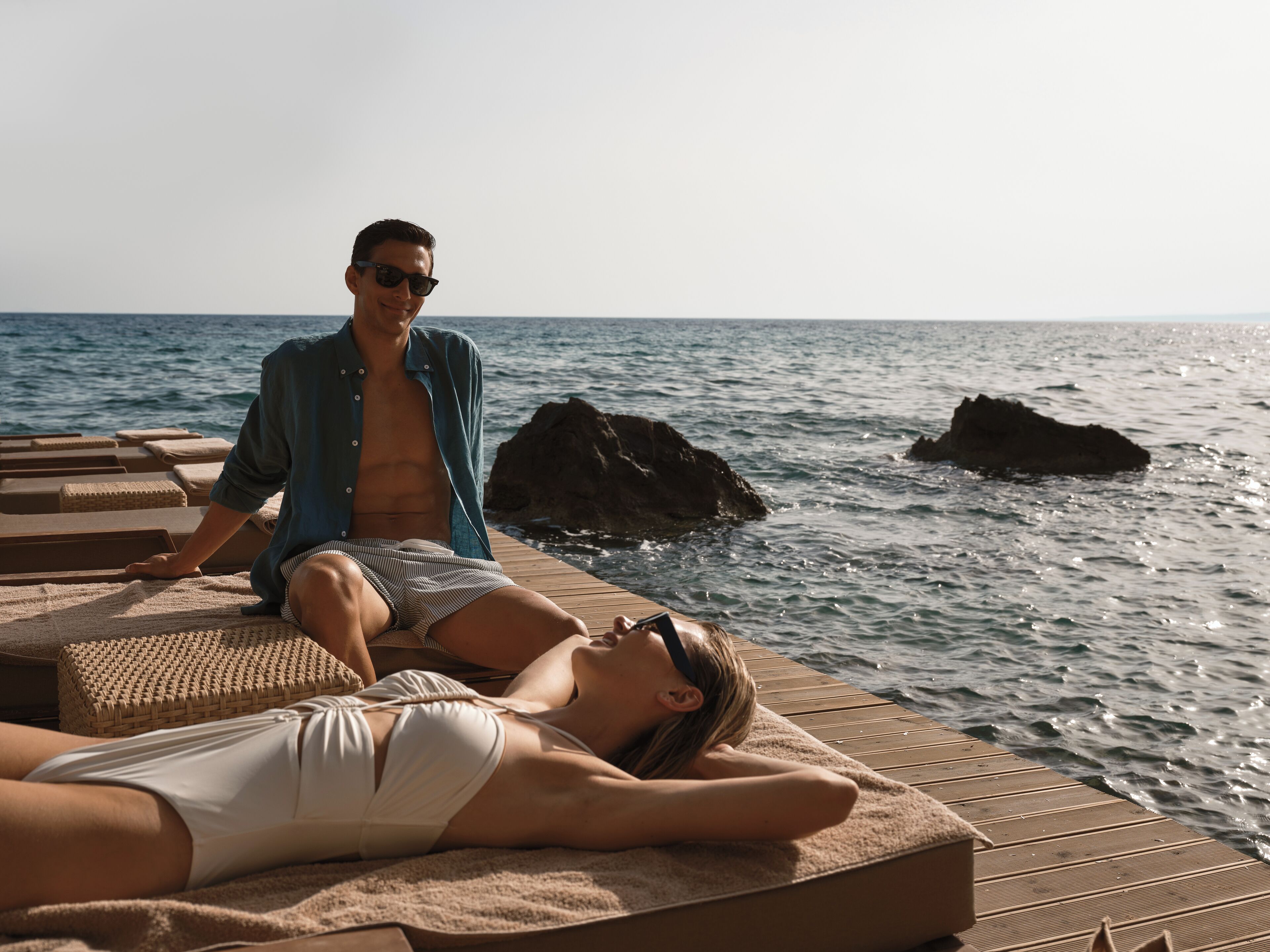 A couple relaxing on a wooden deck with a view of the sea and rocks.