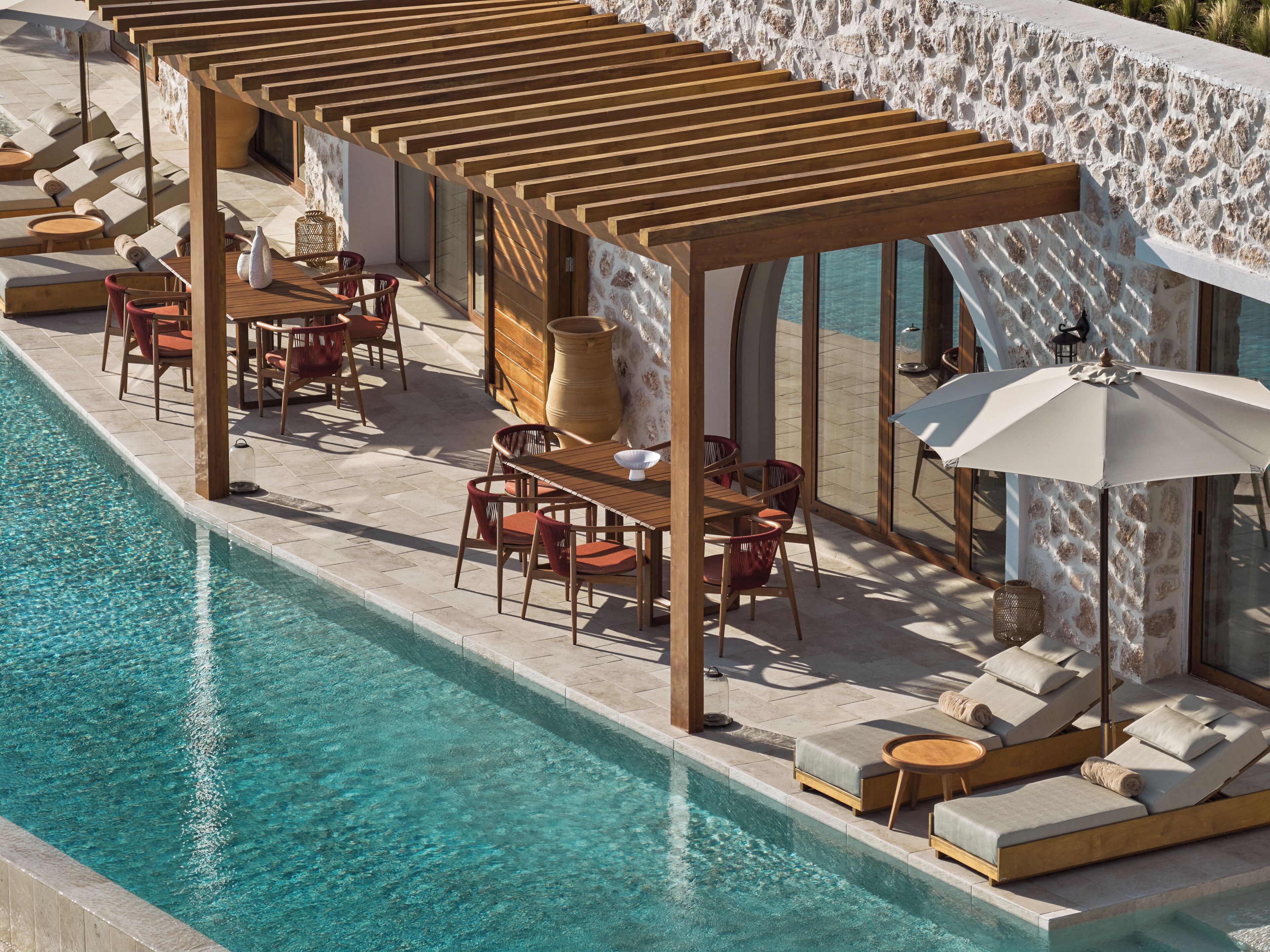Een zwembadgedeelte met houten pergola’s, ligbedden en een tafel met stoelen.