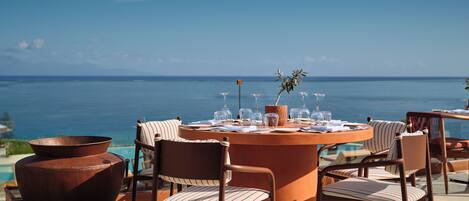 A table set for dining with chairs, a potted plant, and a view of the sea.
