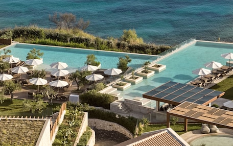 A large outdoor swimming pool with multiple sun loungers and umbrellas, surrounded by greenery and overlooking a body of water.