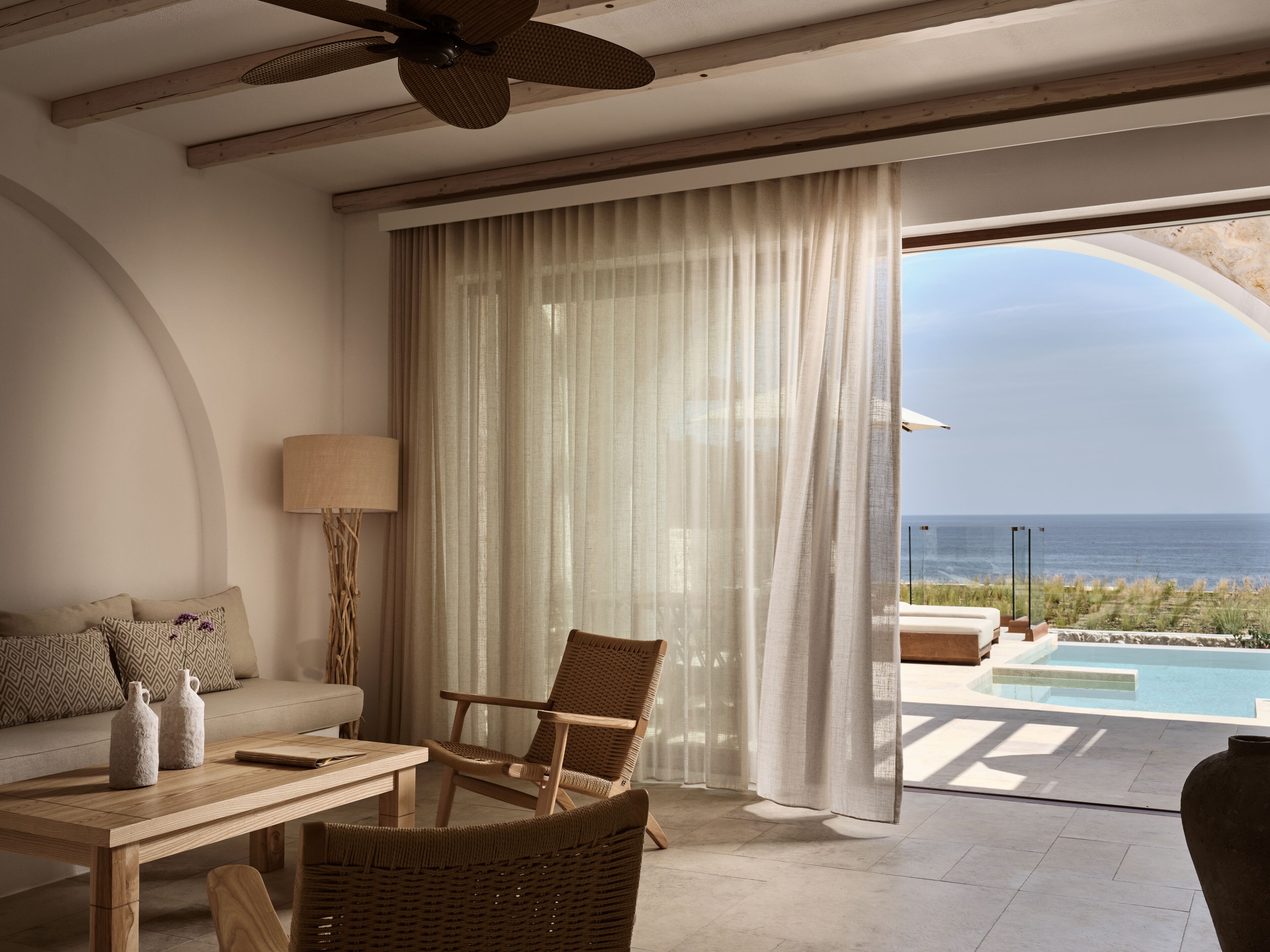 A room with a wooden ceiling, a ceiling fan, a sofa with patterned cushions, a dining table with two bottles, a wicker chair, and a view of the ocean through large curtains.