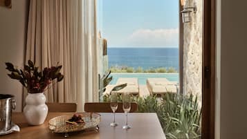 A dining table with a vase of dried flowers, a fruit bowl, and two wine glasses, overlooking a pool and ocean view.