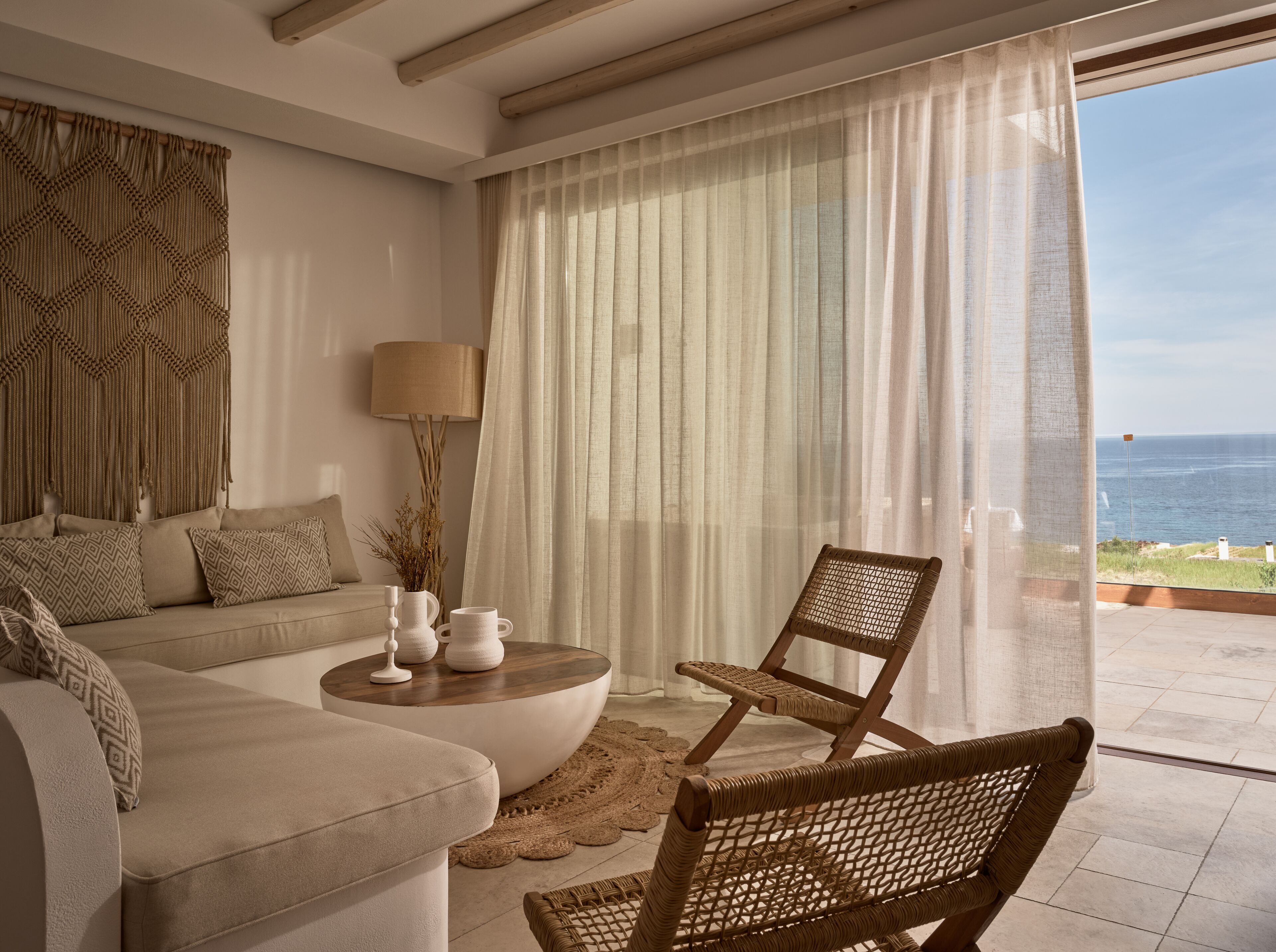 Un salon comprenant un canapé, une table basse ronde, un fauteuil en osier et une vue sur la mer, visible à travers de fines rideaux.