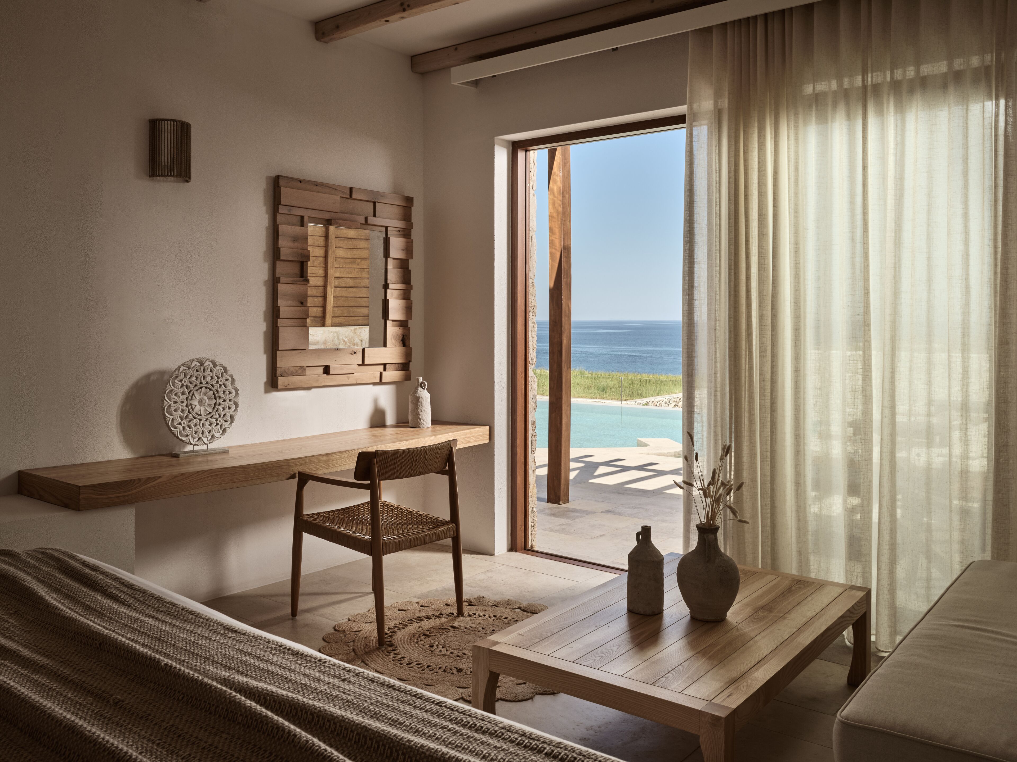 A room with a wooden desk, a wicker chair, a small table with vases, a wall-mounted decorative piece, and a view of the ocean through a glass door.