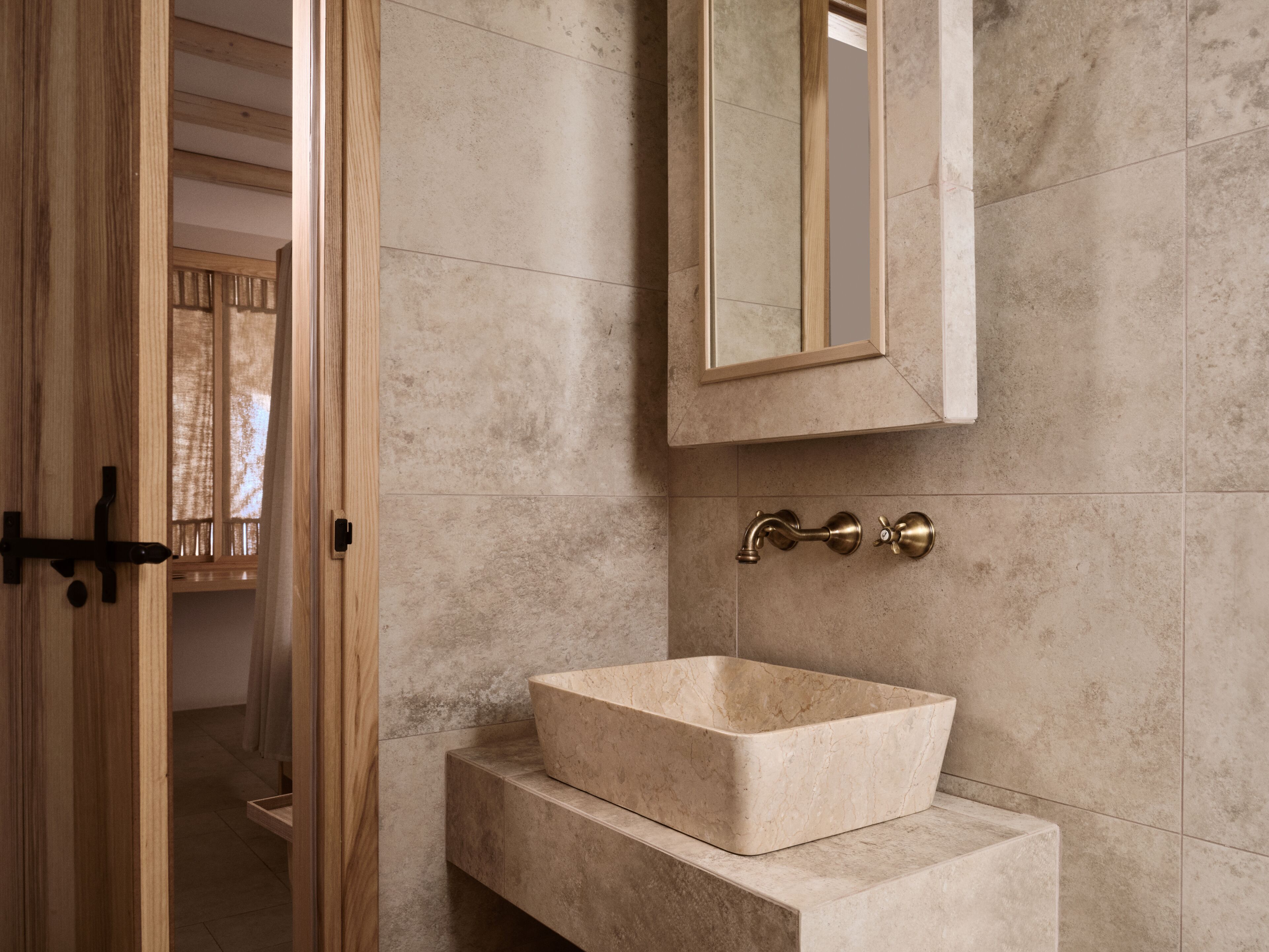 Un baño con puerta de madera, un lavabo de mármol color beige y un espejo instalado en la pared.