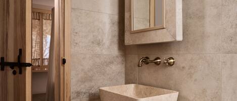 Un baño con puerta de madera, un lavabo de mármol color beige y un espejo instalado en la pared.
