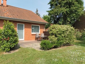 Une maison en briques avec un toit en tuiles, une terrasse avec une table et des chaises, et un jardin agrémenté de divers arbustes et d’une pelouse.