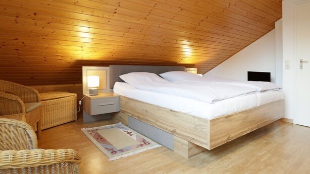 A bedroom with a wooden ceiling, a bed with white bedding, a nightstand with a lamp, and wicker furniture. - Urlauberdorf 50a, Deutschland (Ostseebad Boltenhagen)