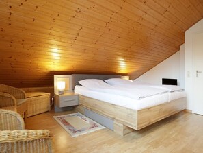 A bedroom with a wooden ceiling, a bed with white bedding, a nightstand with a lamp, and wicker furniture.
