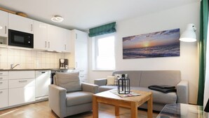 A modern living room with a grey sofa, a wooden coffee table, and a kitchen area with white cabinets and a microwave. - Urlauberdorf 50a, Deutschland (Ostseebad Boltenhagen)