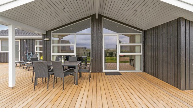 Een overdekt buitengebied met een houten terras, een tafel met stoelen en grote schuifdeuren die uitkomen op het uitzicht op de natuur.