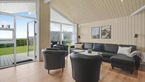 Een ruime woonkamer met een grote schuifpui naar een buitenruimte, een donkere bank en een houten plafond.