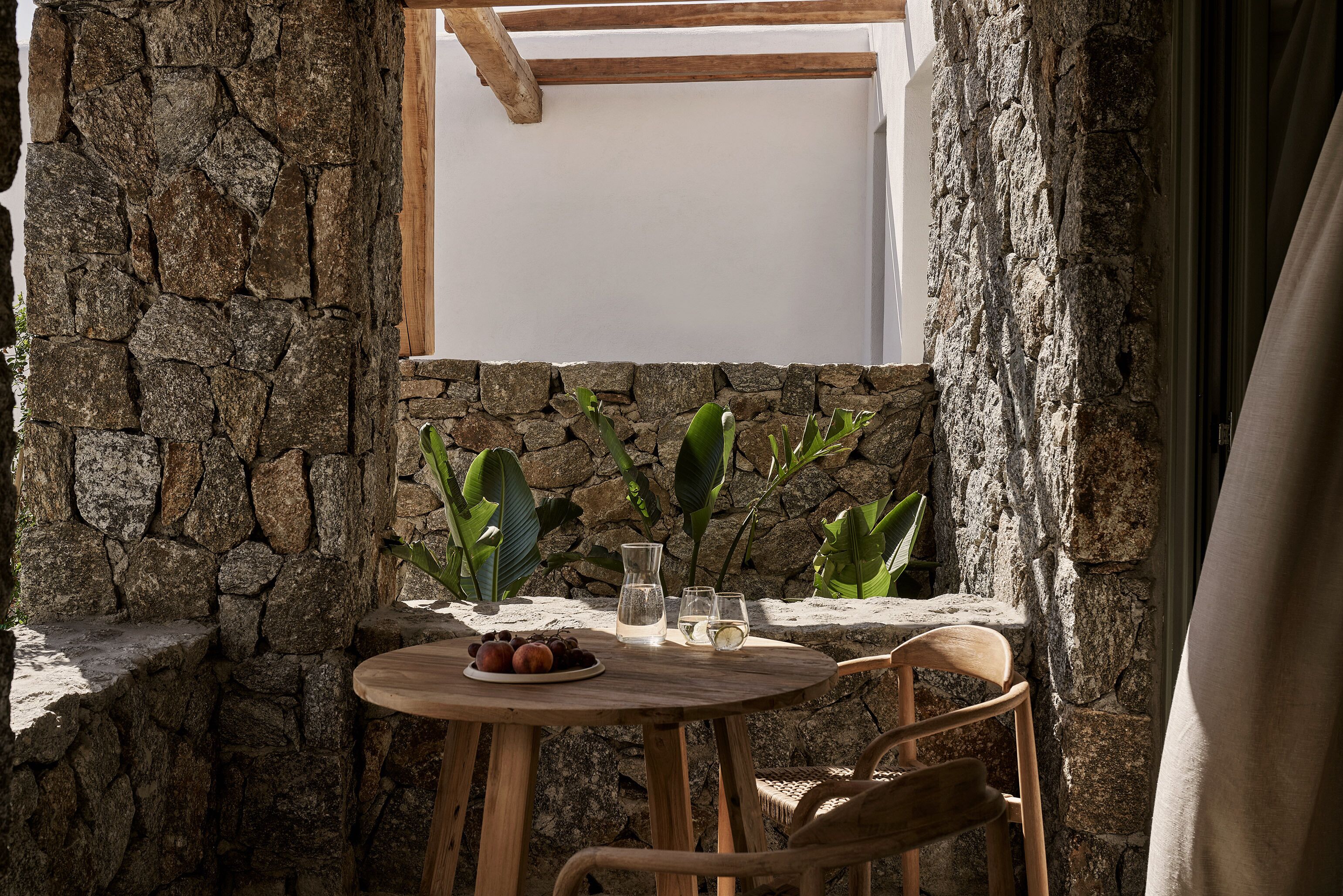Un mur de pierre, une table en bois avec une assiette de fruits, deux verres et une carafe.