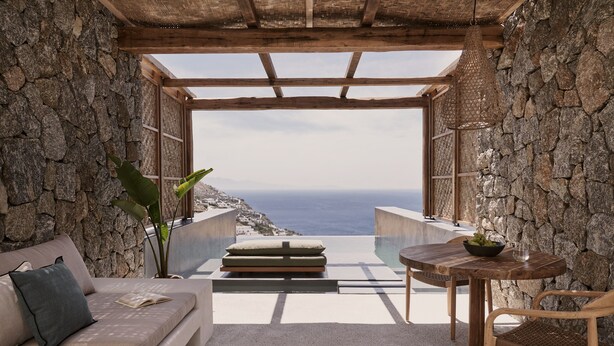 Una habitación de paredes de piedra con techo de madera, un sofá, una mesa con un cuenco de fruta y vistas al mar.