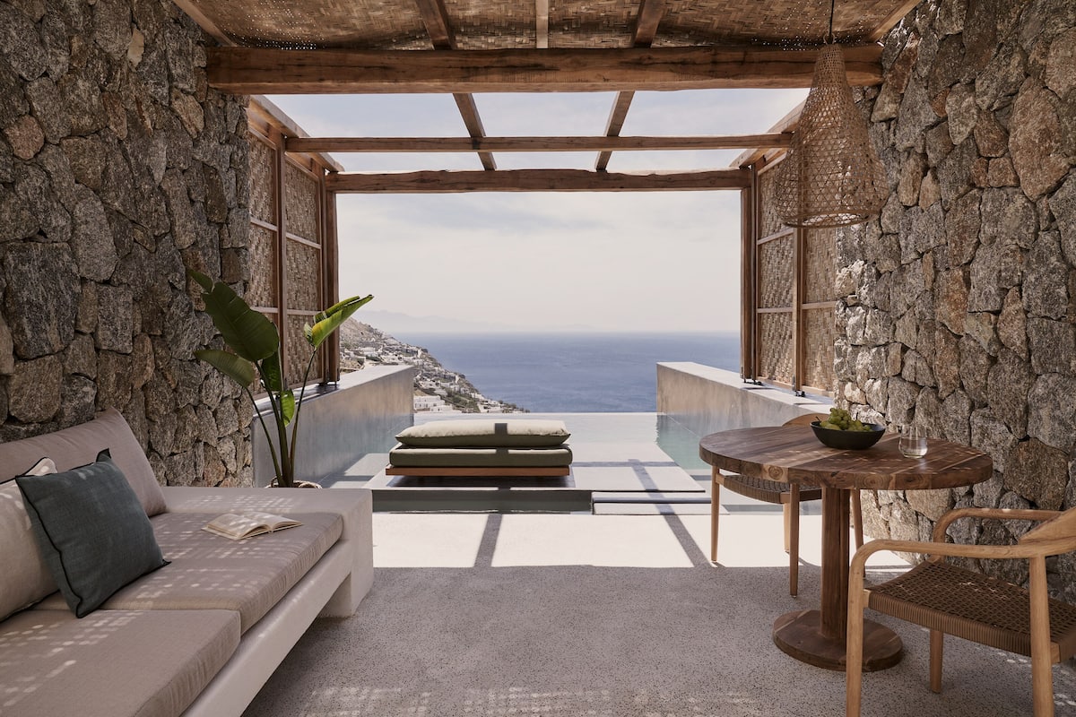 Quarto com paredes de pedra e teto de madeira, um sofá, uma mesa com uma tigela de frutas e vista para o mar.