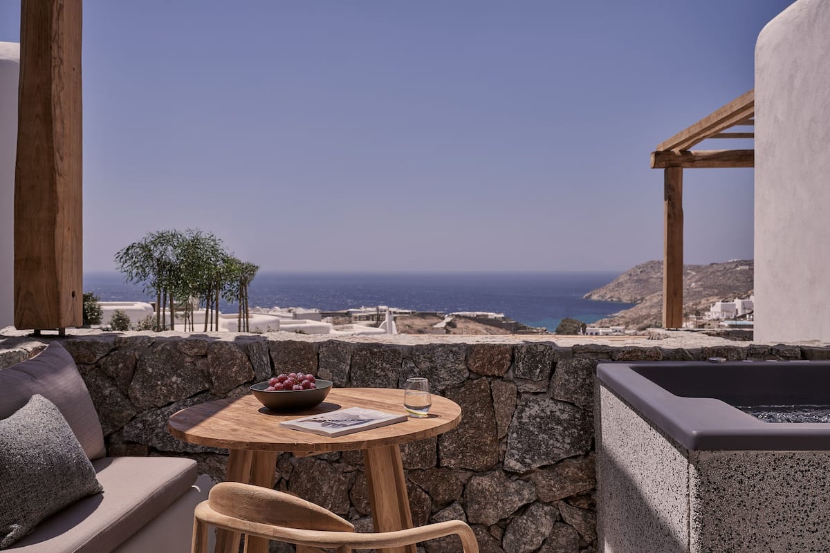 Um muro de pedra, uma mesa de madeira com uma tigela de frutas, um copo de água e vista para o mar.