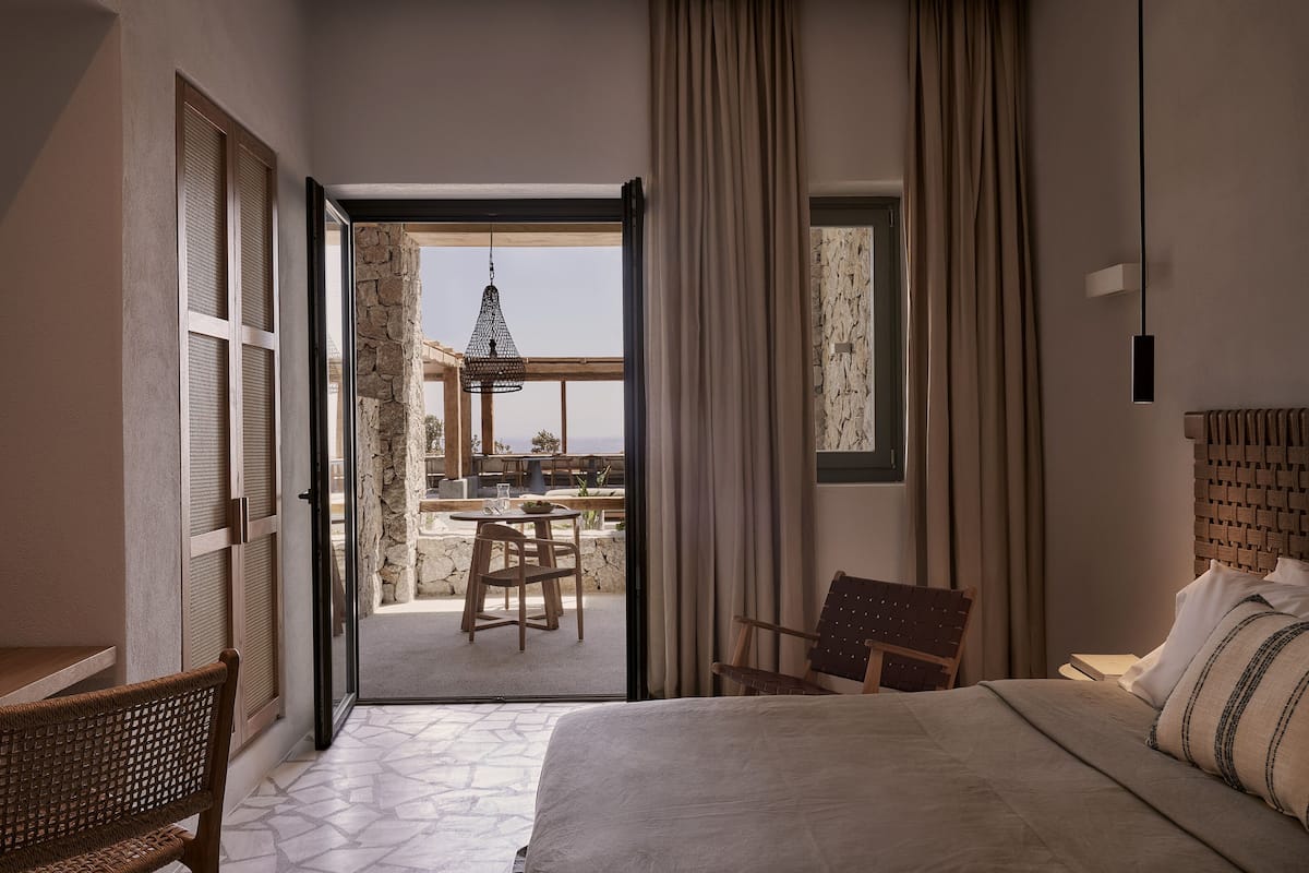 Quarto de hotel com cama, poltrona, mesa e vista para área externa com mesa e cadeiras.