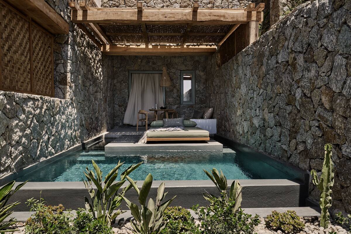 Quarto com paredes de pedra, piscina integrada, cama e uma pequena mesa com cadeira.