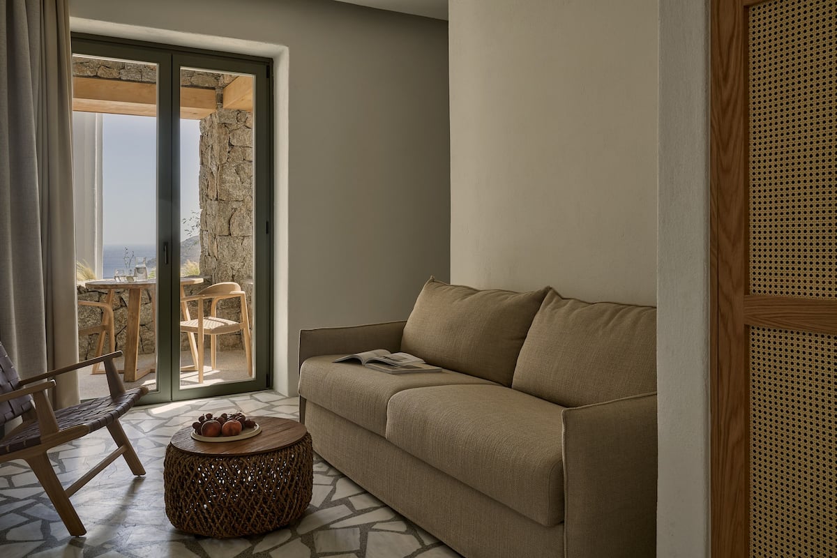 Uma sala de estar com sofá, uma cadeira de madeira, uma mesa redonda pequena com frutas, e vista através de uma porta de vidro para uma área externa com mesa e cadeiras.