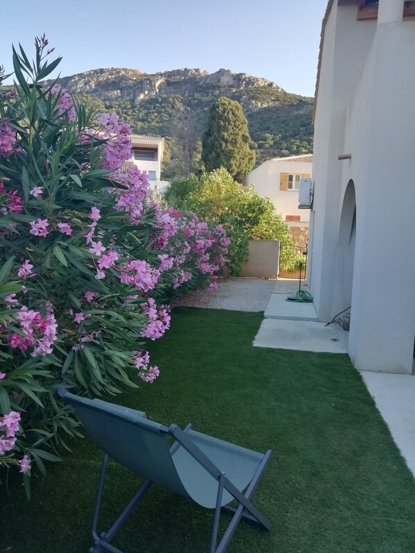 A garden with artificial grass, a folding chair, and a white building with a mountain backdrop. - Villa DUPLEX AVEC JARDIN VUE MER (algajola)